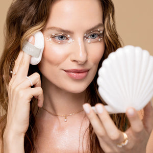 Woman using a white pearl puff and holding a round shell mirror compact