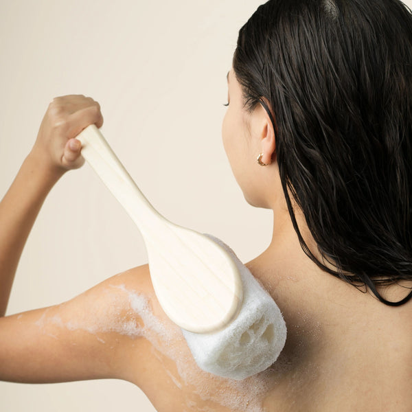 Loofah bath brush scrubbing an arm with white lather and visible foam
