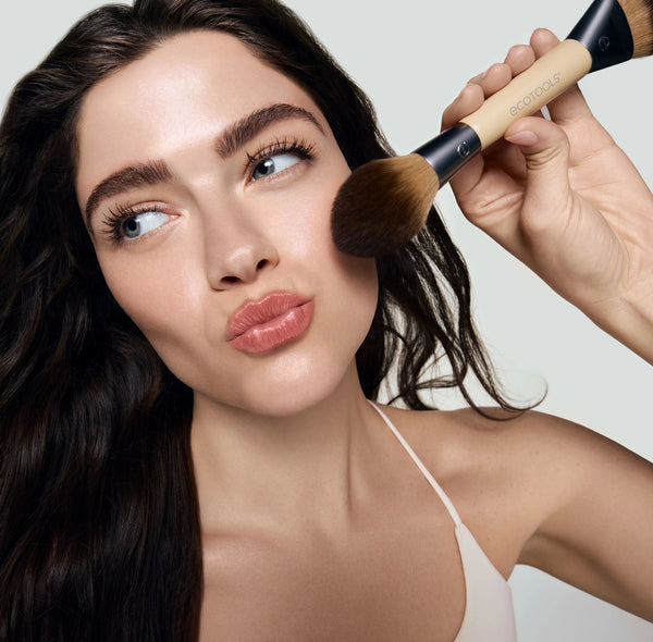 Makeup brush with bronze bristle tip held near face; beige and gray handle