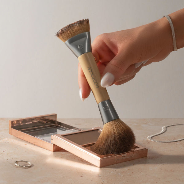 Hand holding dual-ended makeup brush with bronze bristles and gray-beige handle