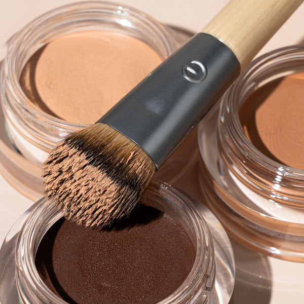Precision concealer brush with bristles coated in brown powder over open jars