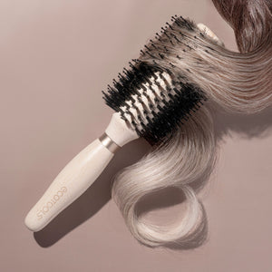 Volumizing round brush brushing through light blonde hair; light wooden handle and black bristles