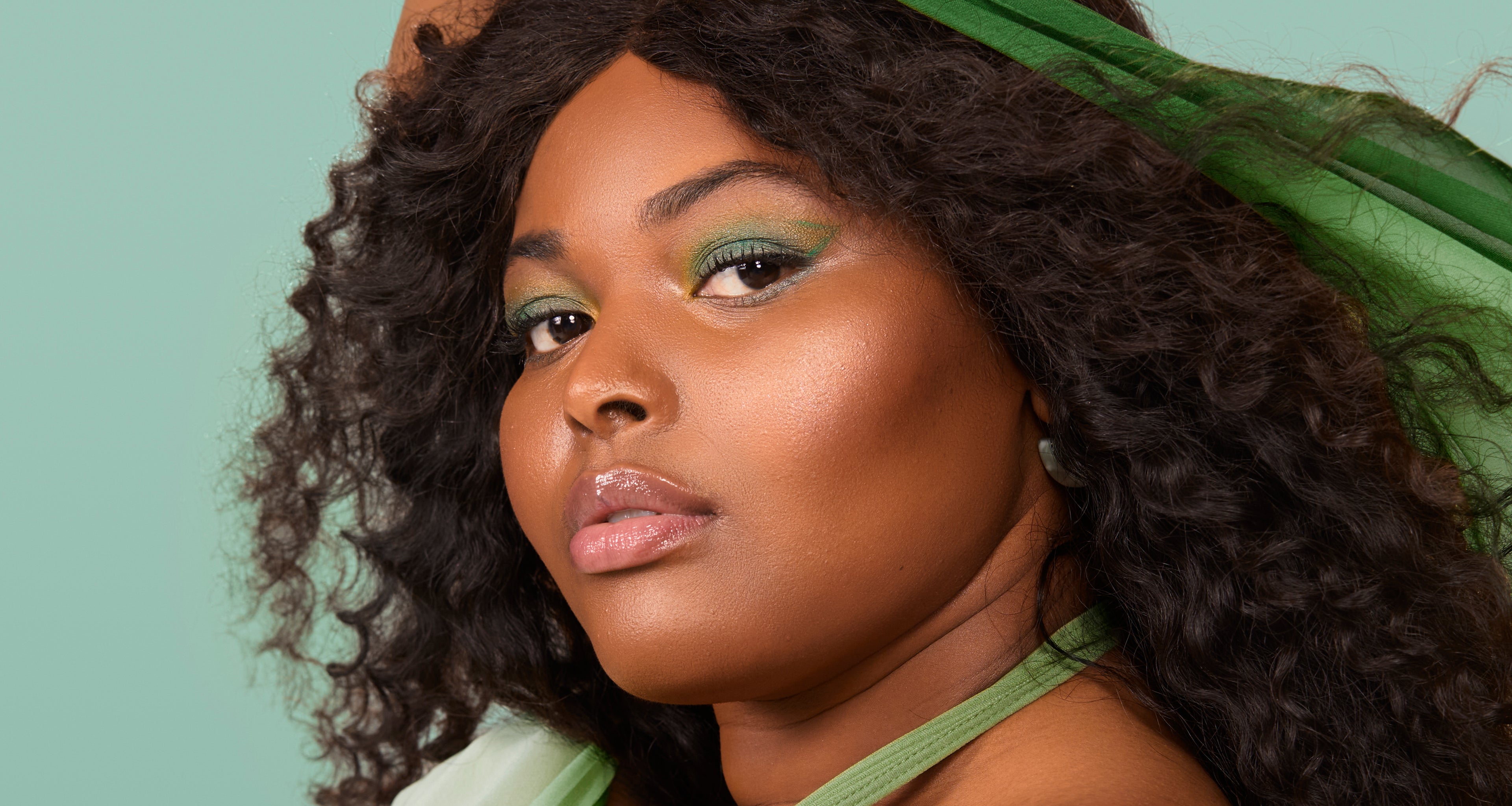 Close up of a woman with dark curly hair, green headband, and green eye makeup against a light teal background