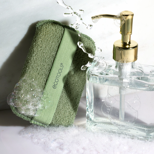 Olive-green exfoliating body buffer with strap beside soap bubbles