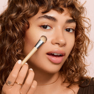 Precision concealer brush used on under-eye area by a person with curly hair