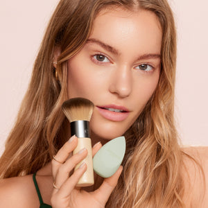 Facial makeup brush and mint blending sponge held by a person