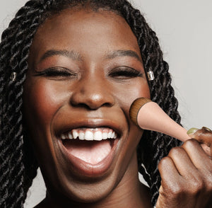 Makeup brush held against cheek, with round golden bristle head and pink handle