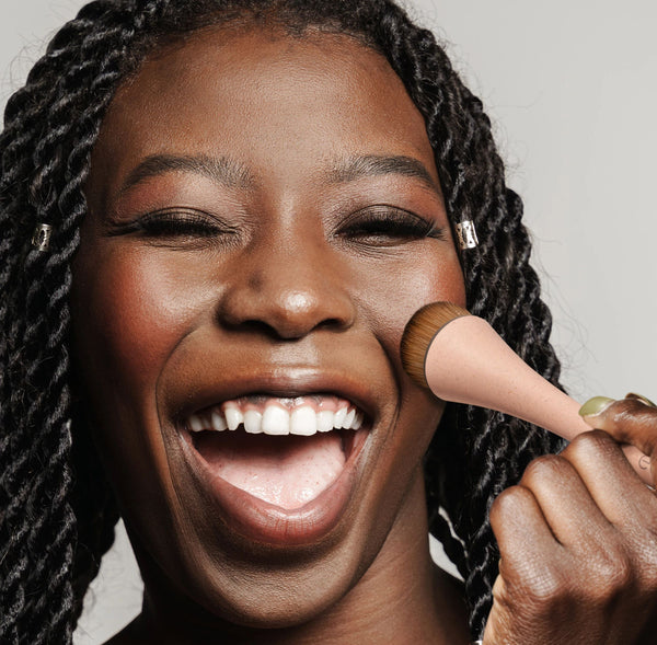 Makeup brush held against cheek, with round golden bristle head and pink handle