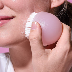 Pink facial brush with white bristles being used on cheek by hand