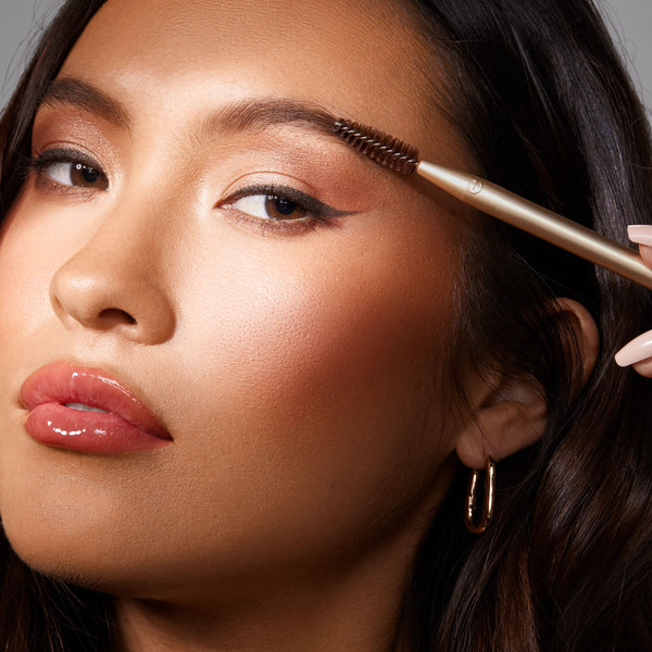 Woman applying eye makeup with a gold brush across the eyelid; spoolie tip visible.