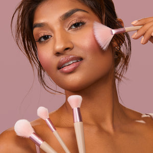 Makeup brush being used to apply blush; pink-bristled brush with beige handle near cheek.