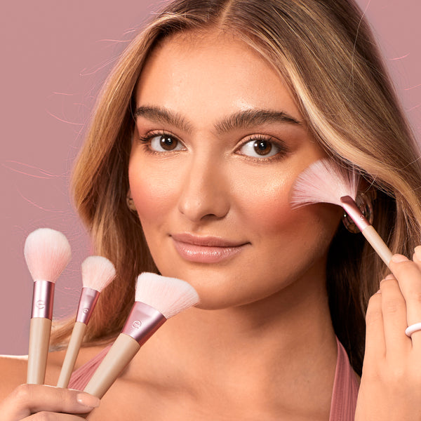 Woman holding pink-bristled makeup brush with beige handle near temple, using the brush.