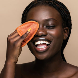 Woman smiling with orange teardrop cleansing cloth on cheek for gentle facial cleansing