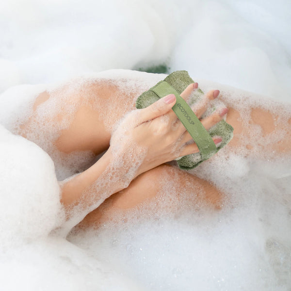 Olive-green exfoliating body buffer being used with lather and bath bubbles