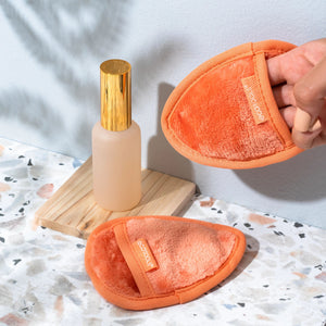 Orange facial cleansing cloth held on counter next to cleanser bottle