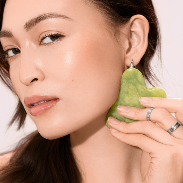 Woman holds jade gua sha while using jade facial roller on her cheek