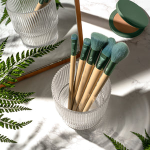 Teal-and-beige face makeup brushes in a clear glass container with greenery nearby