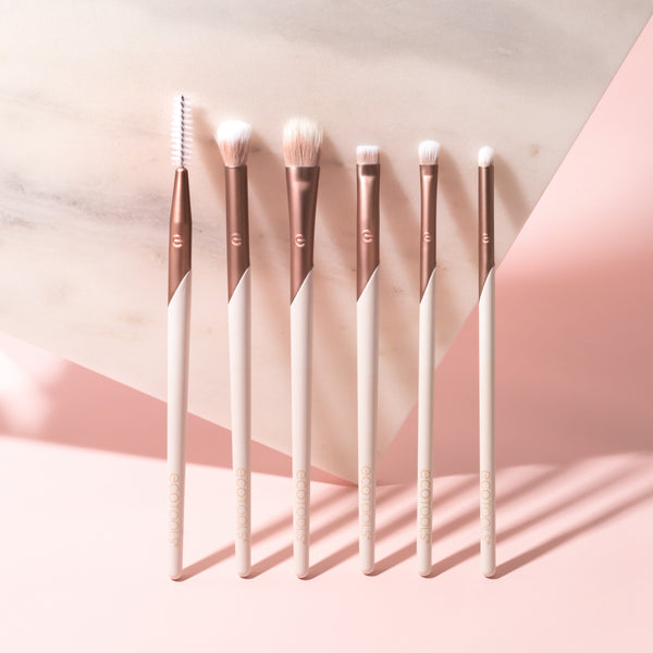 Seven rose-gold eye brushes with white bristles arranged in a row, including spoolie