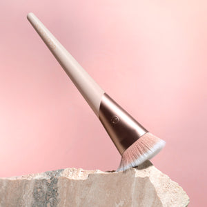 Angled foundation brush with soft bristles, rose-gold collar, beige handle on stone