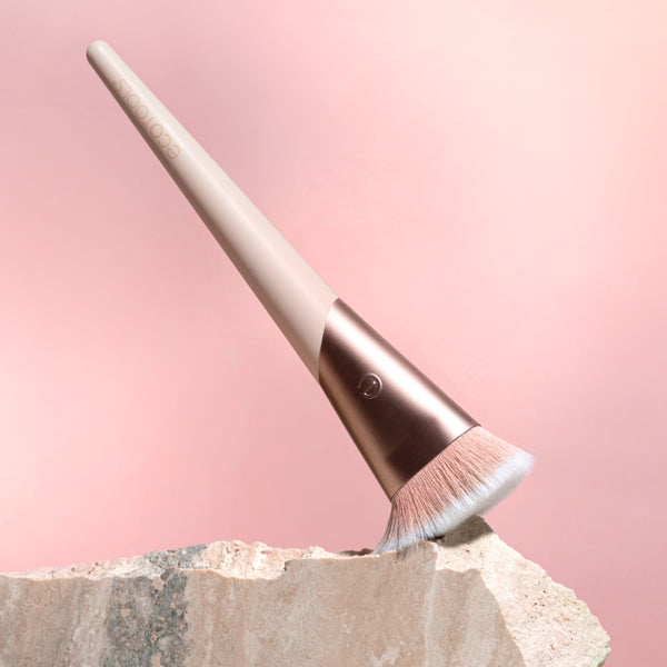 Angled foundation brush with soft bristles, rose-gold collar, beige handle on stone