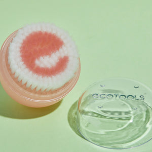 Pink facial cleansing brush head with dense bristles, beside clear brush cover